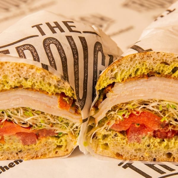 A turkey sandwich cut in half, wrapped in branded paper, revealing layers of meat, sprouts, and tomato.