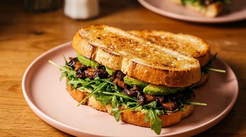Toasted bread sandwich with avocado slices, mushrooms, and arugula on a pink plate.