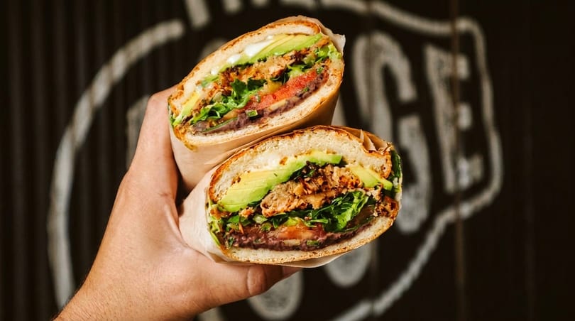 A hand holds two halves of a large sandwich filled with meat, avocado, lettuce, and tomato.