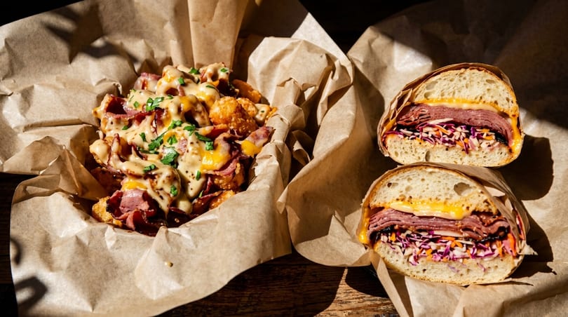 Pastrami tater tots with cheese sauce and two pastrami sandwiches with coleslaw on a wooden table.