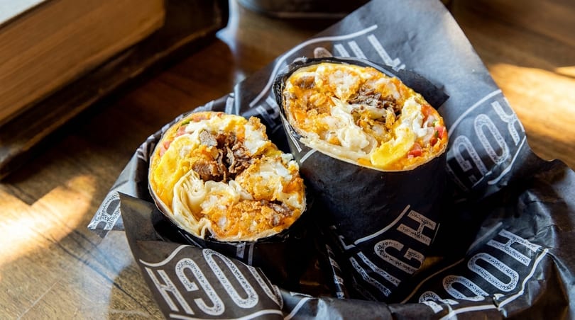 Two halves of a breakfast burrito filled with eggs, meat, and cheese, wrapped in branded paper.