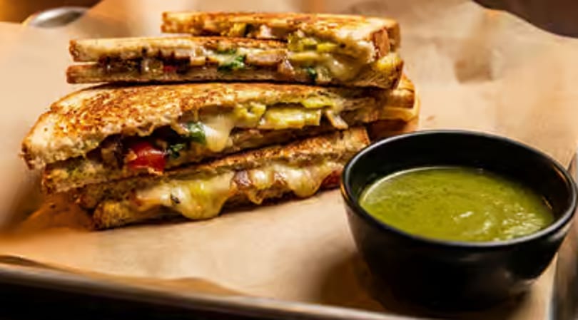 Stacked grilled cheese sandwiches with melted cheese and a side of green dipping sauce.