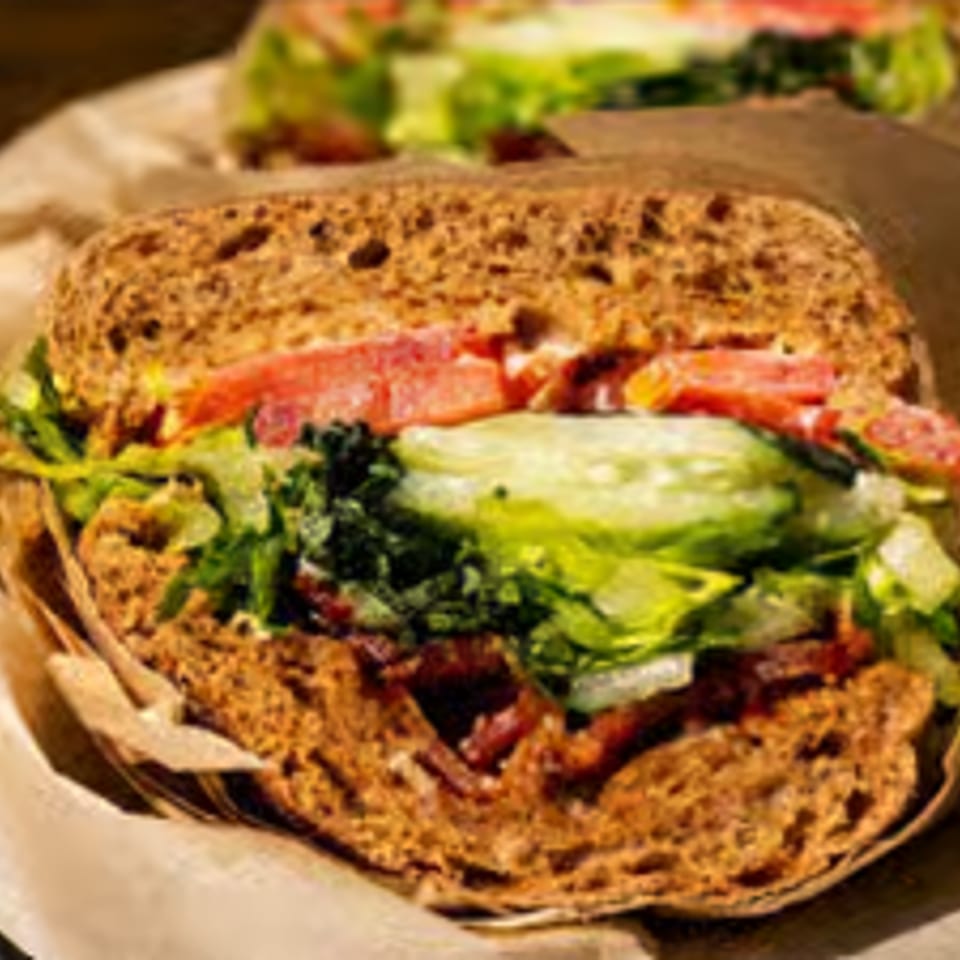 Hearty sandwich with whole wheat bread, lettuce, tomatoes, cucumbers, and bacon on brown paper.